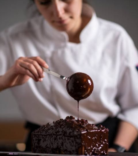 Female baker pouring chocolate sauce on cake with ladle.